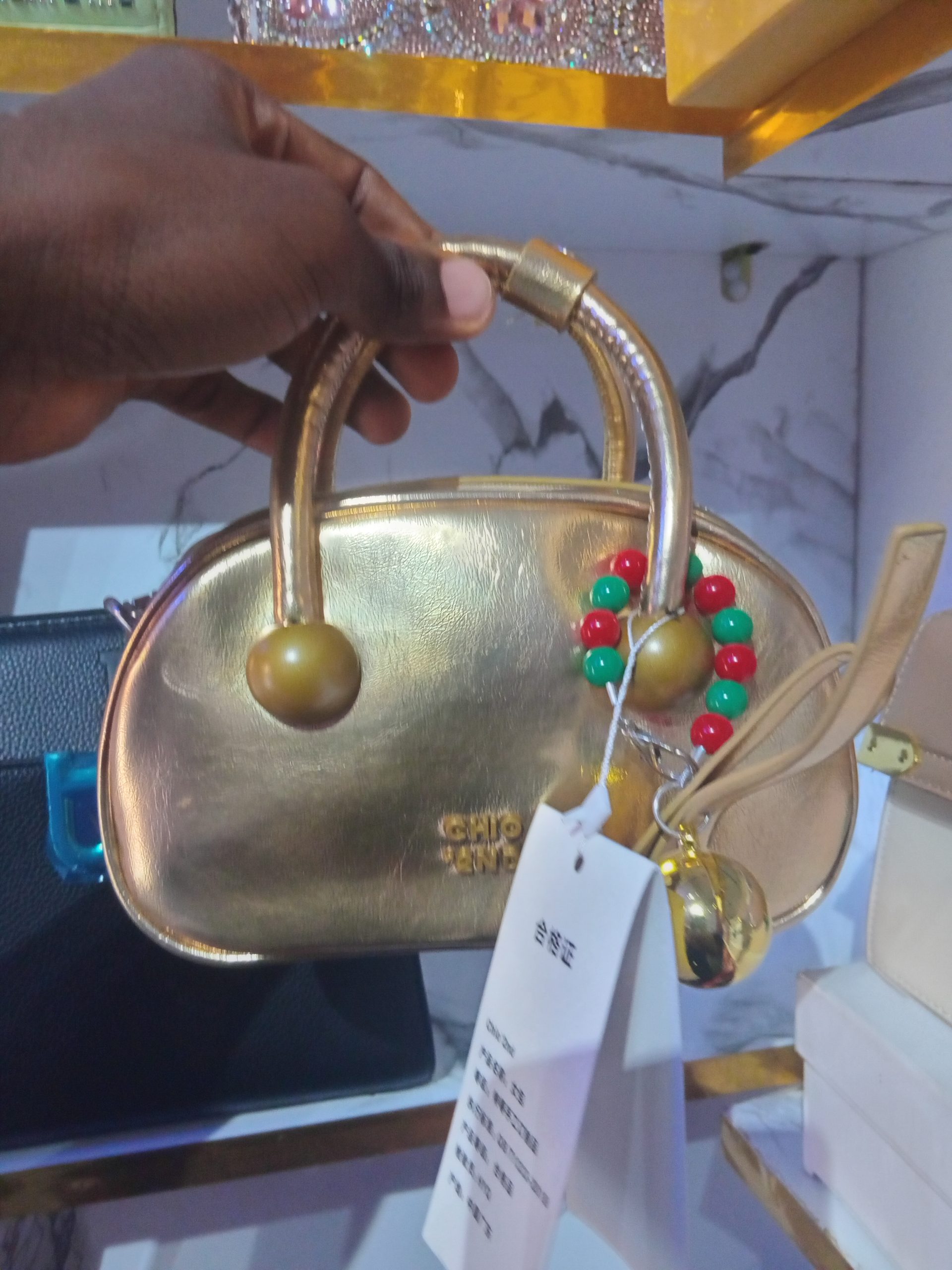 Metallic gold Chio'2nd mini handbag with curved top handle, spherical end-caps, and red and green beaded charm.