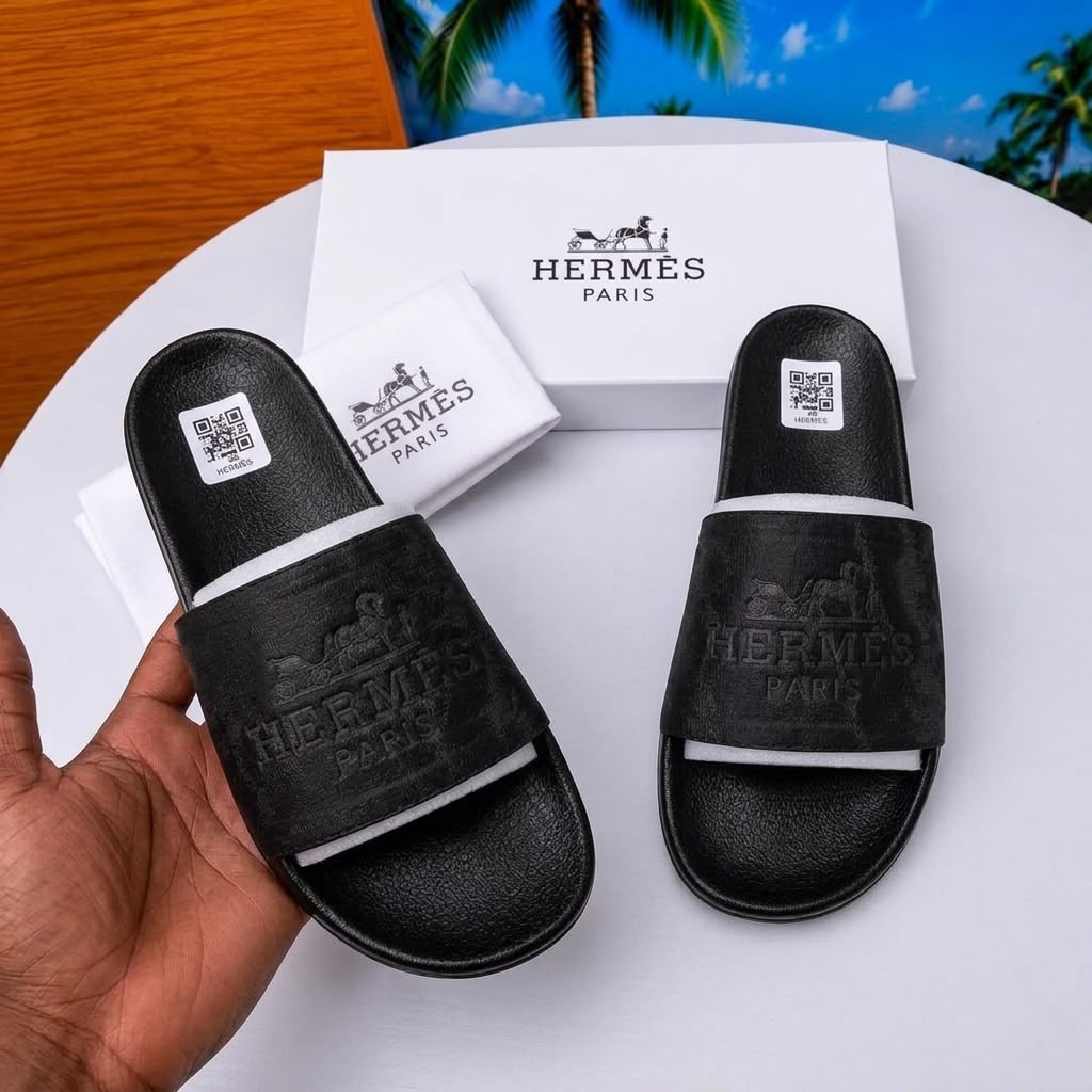 Pair of black Hermès slide sandals with an embossed carriage logo on a white round table beside a white packaging box and dust bag.
