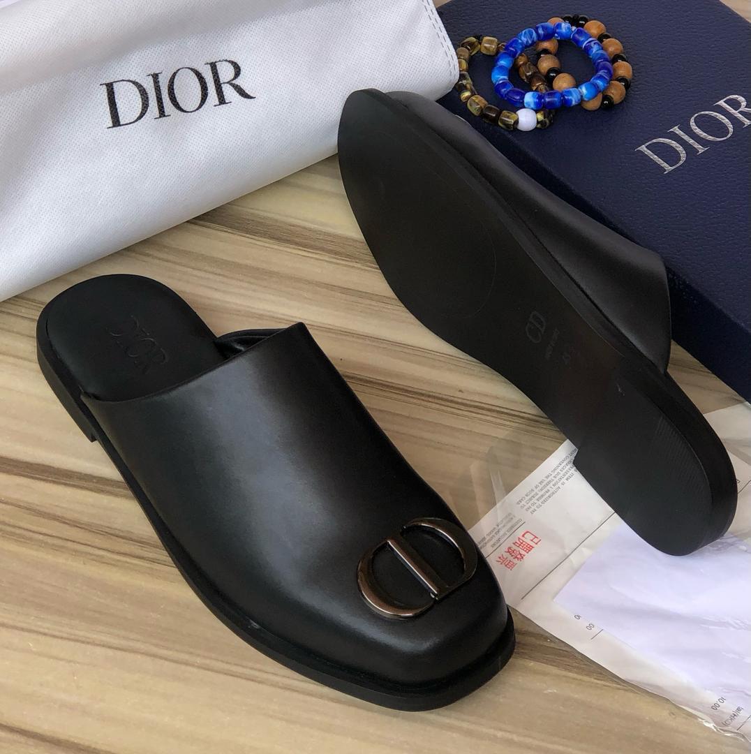 Pair of Dior men's black leather CD buckle mule slippers displayed on a wooden surface next to branded packaging.