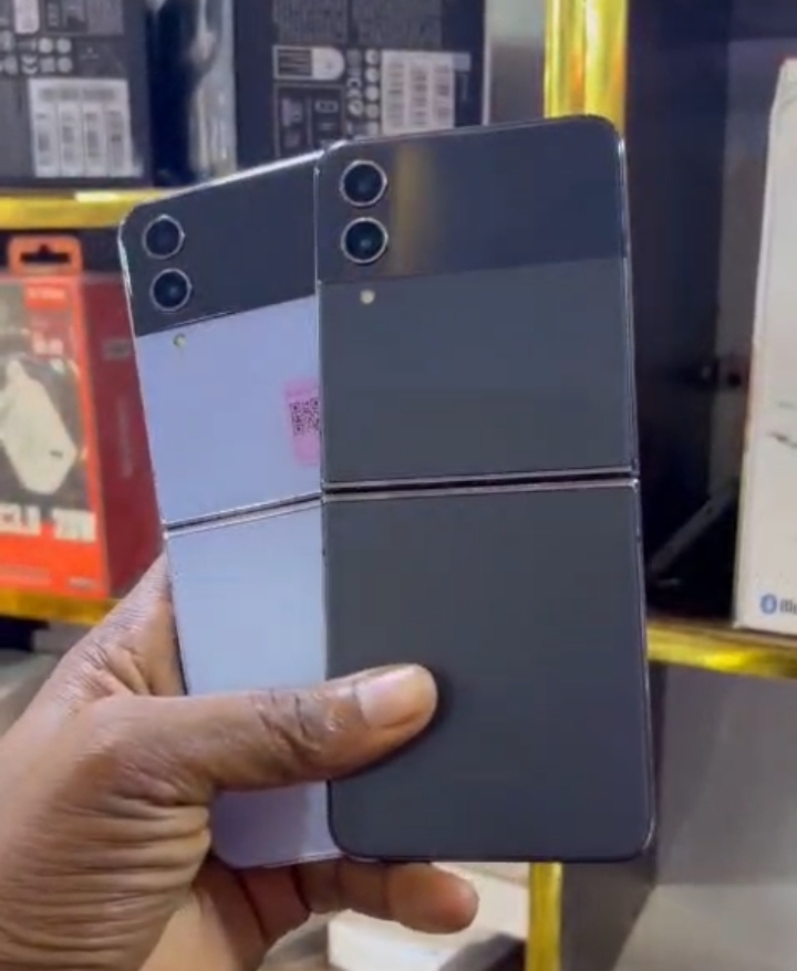 Hand holding two UK Used Samsung Galaxy Z Flip4 smartphones in light purple and dark gray colors.